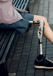 A person wearing shorts sits on a bench, with a prosthetic leg and a sneaker visible. The ground is paved with rectangular tiles.