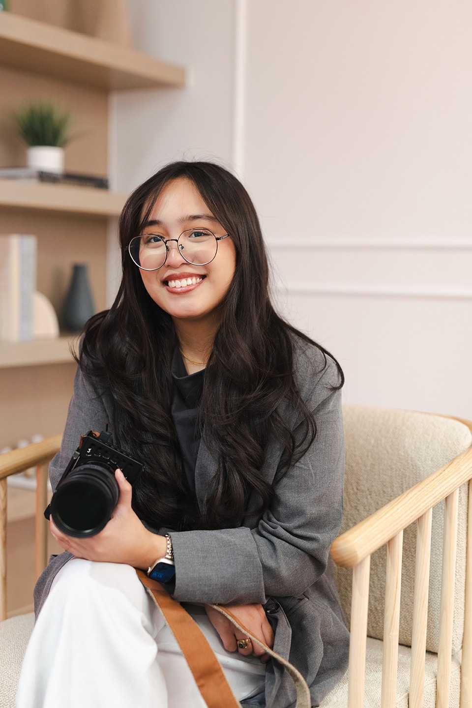 A woman with long dark hair and glasses sits on a chair, smiling, and holding a camera. She is wearing a gray blazer and white pants. Shelves with books and a plant are in the background.